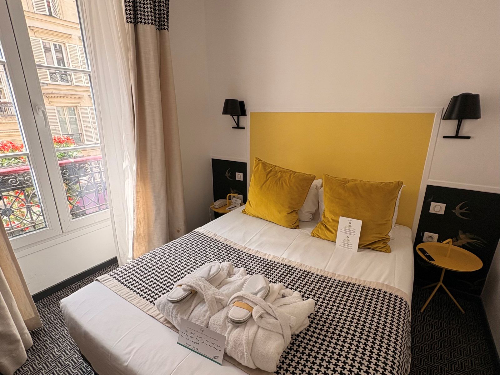 A hotel bed made with robes and slippers placed on top and a window to the side looking out to the street
