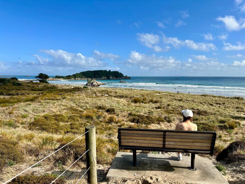 A woman sits on a bench at a beach overlooking the ocean during a sunny morning