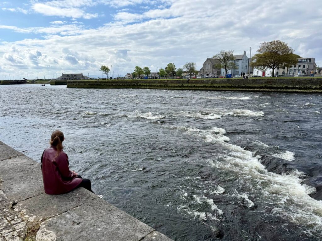 A girl sits looking out at the water in Galway on an Ireland road trip